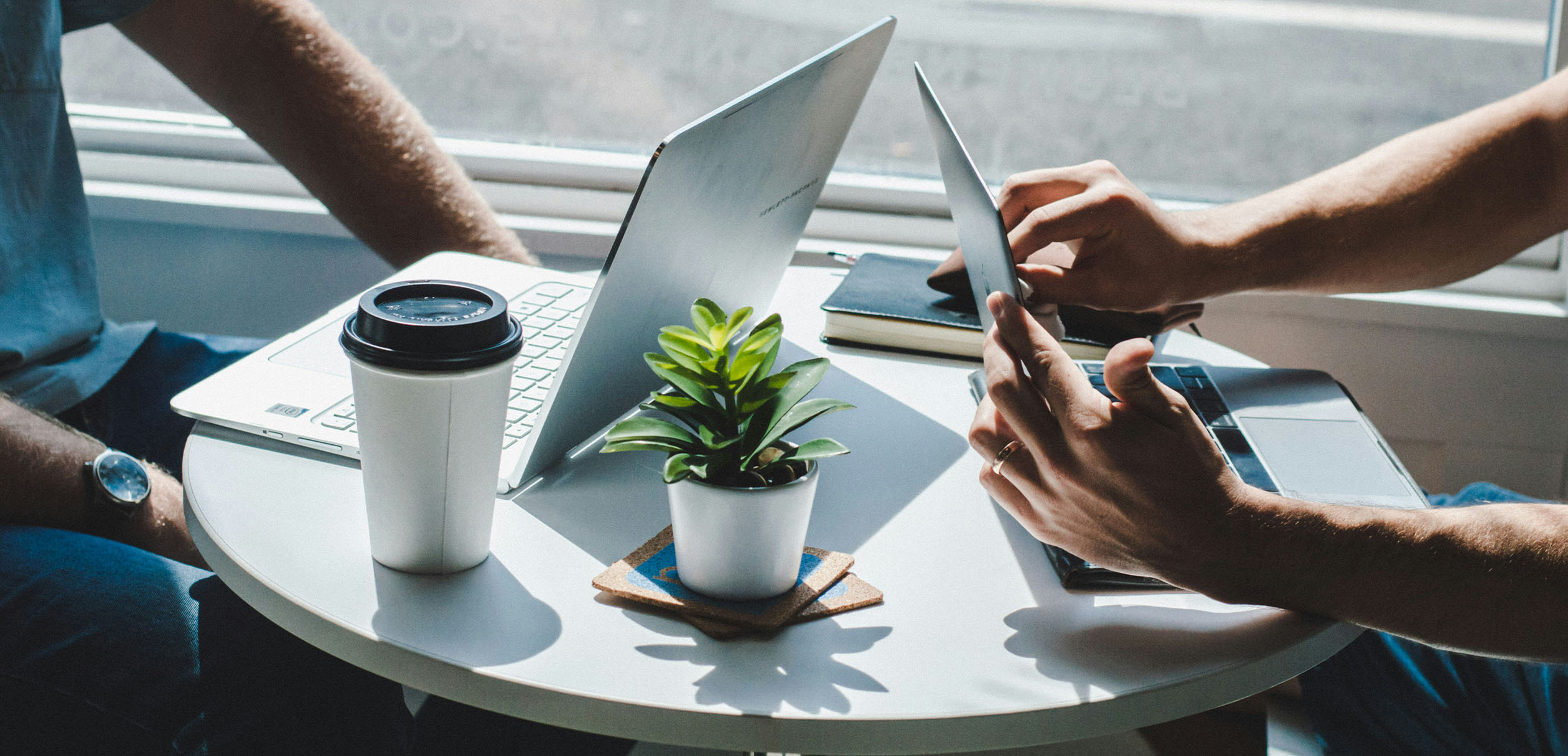 Two people working on laptops at a small round table with a coffee cup, notebook, and potted plant.